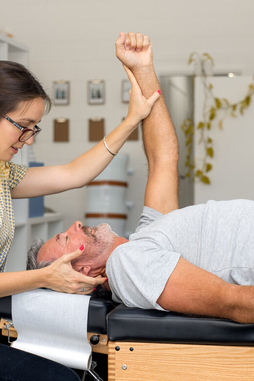 An image of a man laying on a bed as a woman adusts his arm. This could represent how a holistic chiropractor in the Chicago area can offer support with chiropractic care in Orland Park, IL. Learn more about applied kinesiology in Orland Park, IL today.