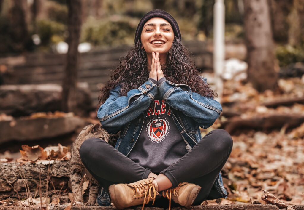 A woman smiles while sitting on a forest floor with her hands together. This could represent the benefits of meditation for hormone levels. Contact a functional medicine doctor in Orland Park, IL to learn more about hormonal weight gain and the benefits of a weight loss program in Orland Park, IL today. 
