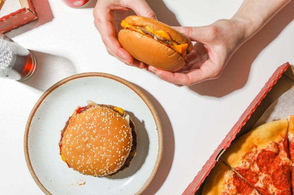 A top down of a person eating a cheeseburger with a pizza nearby. Learn more about how a holistic doctor in Orland Park, IL can offer support with Shape ReClaimed in the Chicago area. Learn more about this weight loss program in Orland Park, IL today. 
