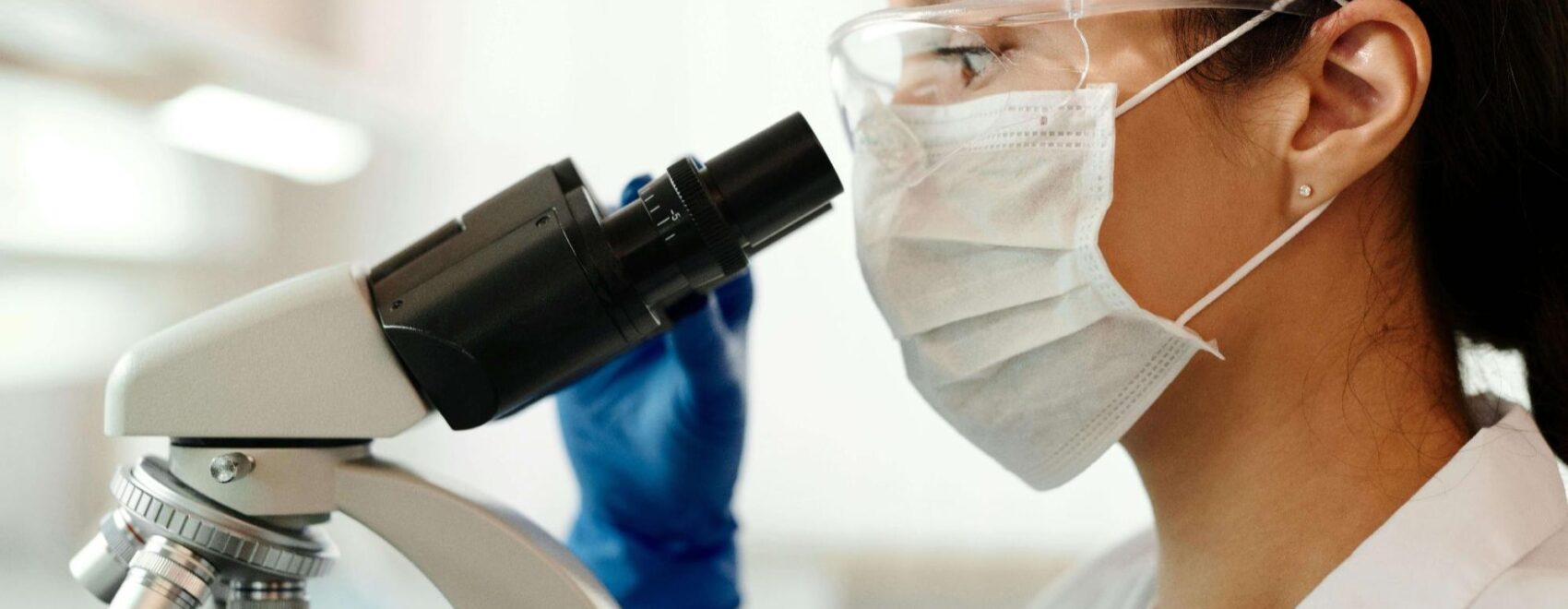 A laboratory scientist wearing protective goggles, a face mask, and gloves examines a sample through a microscope. This could represent the process of hormone testing in Orland Park, IL. Learn more about the effects of a hormone imbalance Orland Park, IL and how a functional medicine doctor in Orland Park, IL can help.