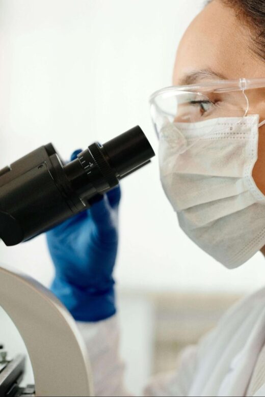 A laboratory scientist wearing protective goggles, a face mask, and gloves examines a sample through a microscope. This could represent the process of hormone testing in Orland Park, IL. Learn more about the effects of a hormone imbalance Orland Park, IL and how a functional medicine doctor in Orland Park, IL can help.