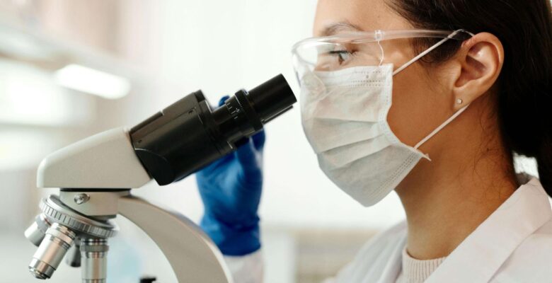 A laboratory scientist wearing protective goggles, a face mask, and gloves examines a sample through a microscope. This could represent the process of hormone testing in Orland Park, IL. Learn more about the effects of a hormone imbalance Orland Park, IL and how a functional medicine doctor in Orland Park, IL can help.