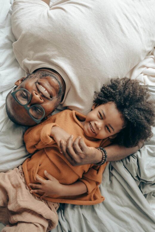 A top down view of a smiling father playing with their child. This could represent the benefits of seeking a therapeutic massage in Orland Park, IL. Search for a massage therapist in Orland Park, IL to learn more about how a holistic massage in Orland Park, IL can help promote oxytocin.