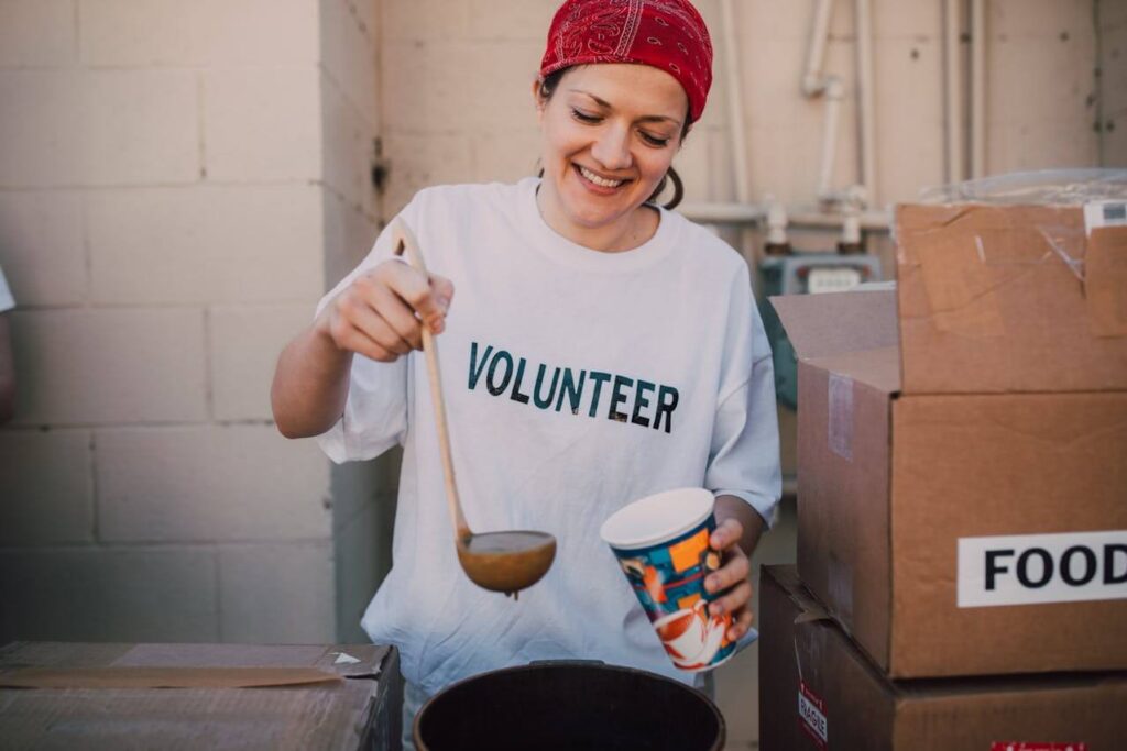 A smiling woman wearing a white "VOLUNTEER" t-shirt and a red bandana is ladling soup into a cup at a food distribution site. Learn how good deeds can promote positive hormones. Search for how a holistic massage in Orland Park, IL can help. Search for a therapeutic massage in Orland Park, IL to learn more.
