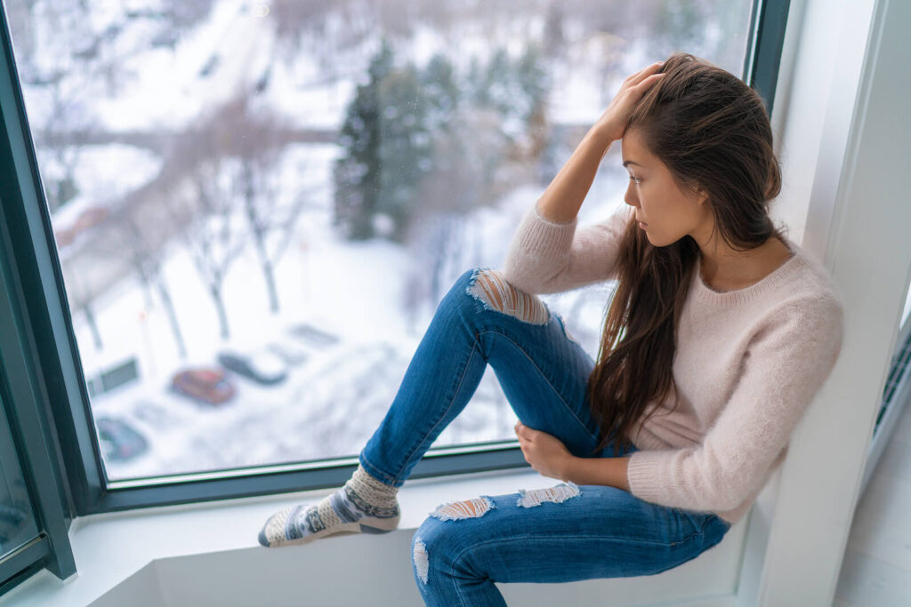 A young woman sits by a large window, gazing outside at a snowy landscape with a thoughtful and concerned expression. This could represent the effects of a hormone imbalance Orland Park, IL. Search for hormone testing in Orland Park, IL to get in contact with a functional medicine doctor in Orland Park, IL today. 
