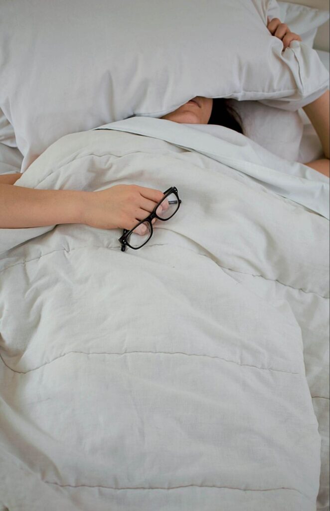 Woman laying in bed holding a pair of glasses and a pillow over her eyes. If you're tired of dragging yourself through the day, find support in restoring your sleep with functional medicine in Orland Park, IL.