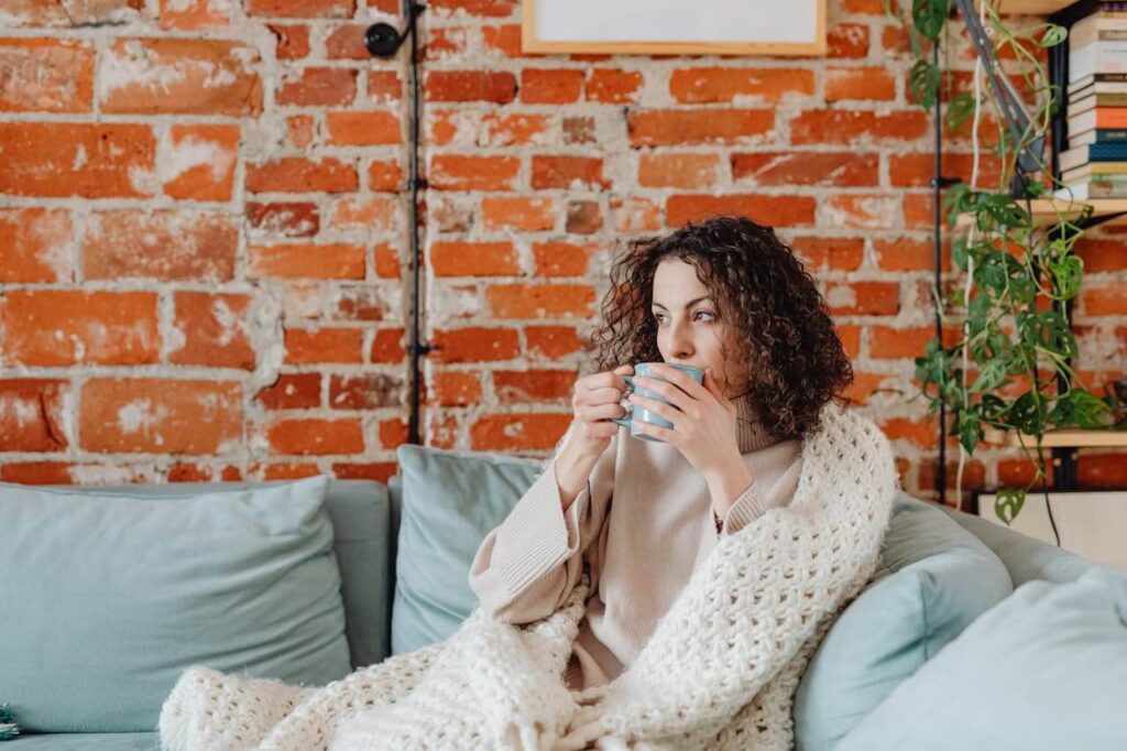 Thoughtful woman sitting on a couch covered with a blanket drinking out of a blue mug. A functional medicine doctor in Orland Park, IL helps uncover the hidden root causes of hormonal imbalance.