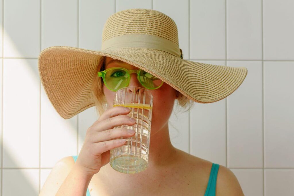green glasses | functional medicine doctor in Orland Park, IL | chiropractic care | chiropractor for sciatica | Homewood | Oak Lawn | Mokena Woman wearing a floppy hat and green sunglasses while drinking a glass of water. From hydration to chiropractic care, functional medicine in Orland Park, IL helps you feel your best all summer long.