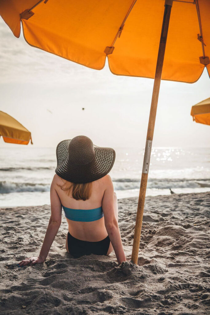 umbrella | functional medicine in Orland Park, IL | chiropractic care | chiropractor | Homewood | Oak Lawn | Mokena Woman sitting on a sandy beach under a yellow umbrella looking at the ocean. Beat summer fatigue and stay active with expert guidance from a functional medicine doctor in Orland Park, IL.