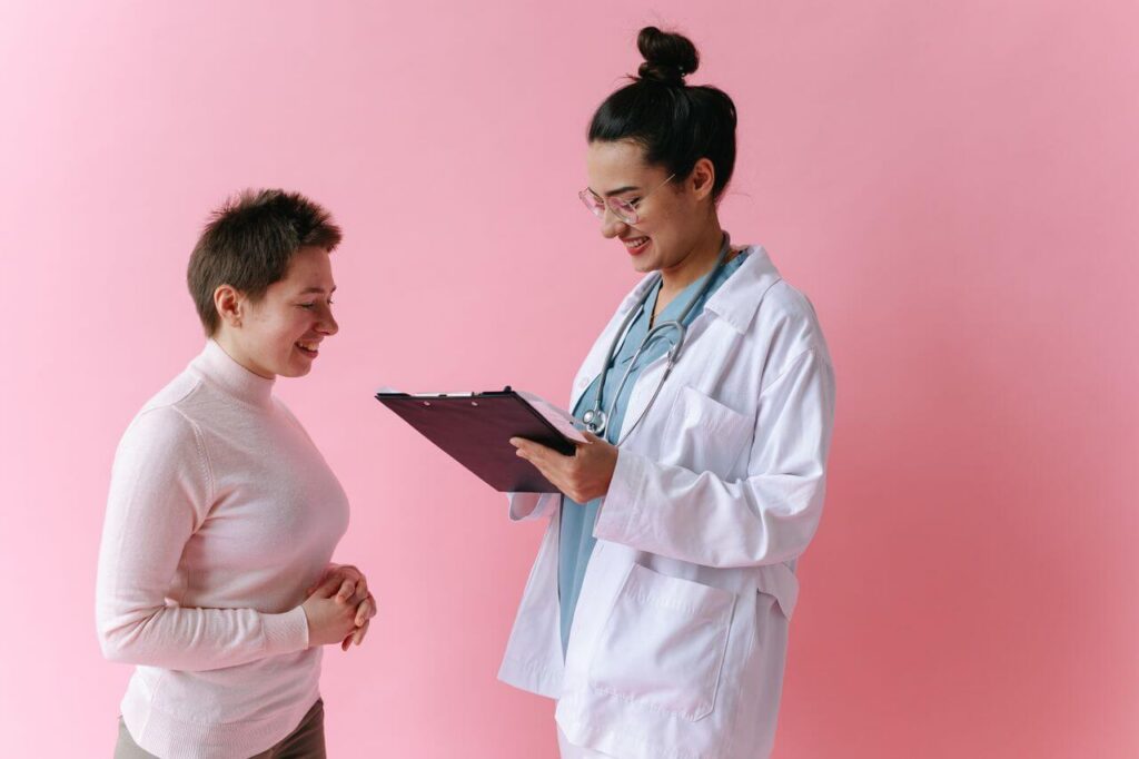 Smiling woman wearing a doctors white coat and stethoscope holding a clipboard and talking to another woman while standing in front of a pink wall. With a supportive a functional medicine doctor, you can discover the root cause behind your PMOS symptoms.