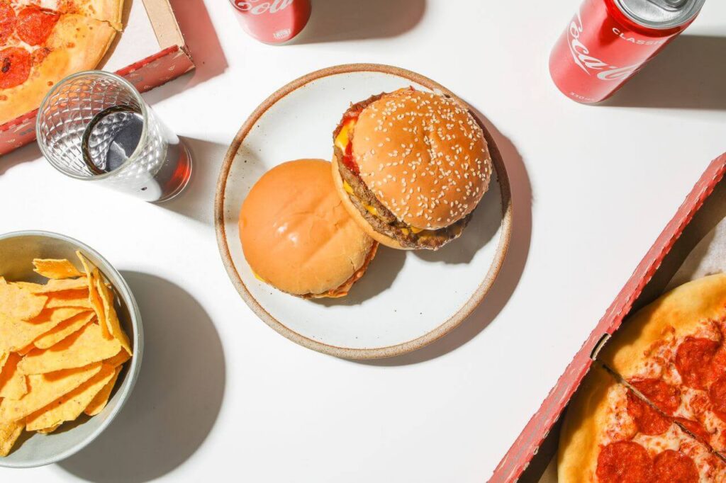 A table with unhealthy fast food burger, chips, pizza and soda.